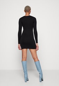 Person med kort hår iklädd en åtsittande svart långärmad miniklänning och blå höga stövlar, stående mot en enfärgad vit bakgrund.