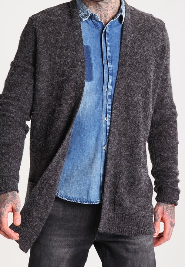 Homme avec des tatouages porte un cardigan ouvert gris foncé sur une chemise en denim bleu et un pantalon noir, debout devant un fond uni.