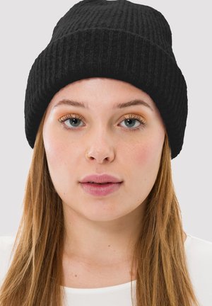 Sort ribbenstrikket hue med en kant, blød tekstur og rund top, båret af en person med lyst brunt hår og et naturligt makeup-look.