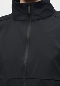 Gros plan d’une personne portant une veste noire avec un col haut et une fermeture éclair centrée, verticale.