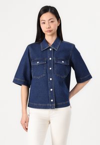 Denim srajca temno modre barve s kratkimi rokavi, zapiranjem na gumbe, dvema žepoma na prsih in podrobnostmi v kontrastnem šivanju. Gladka tekstura.