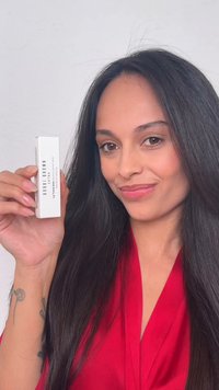 Femme aux longs cheveux foncés et top rouge tenant une boîte de baume à lèvres teinté Bobbi Brown, souriante sur un fond clair uni.