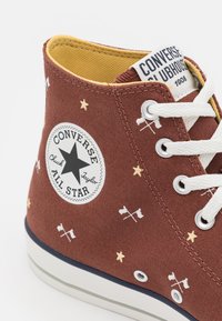 Brązowy wysokotopowy sneaker z płótna, z białymi sznurówkami, wzorami w gwiazdy i flagi, z czarnym logo gwiazdy i żółtą wyściółką.