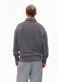 Grijze sweatshirt met een kraag, ribgebreide boorden en zoom. Zachte textuur, ontspannen pasvorm. Draagt met lichtgrijze joggingsbroek, zichtbaar zakdetail.