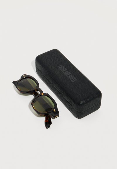 Γυαλιά ηλίου με ταρταρούγα και πράσινους φακούς, τοποθετημένα δίπλα σε μια μαύρη υφή ορθογώνια θήκη γυαλιών ηλίου πάνω σε λευκή επιφάνεια.