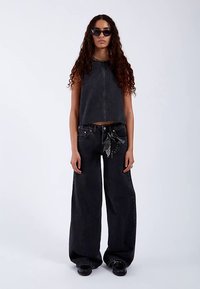 Svart croptopp i denim med en rak halsringning, kombinerad med högmidjade svarta vida jeans med kedjedetalj. Accessoarer inkluderar solglasögon.