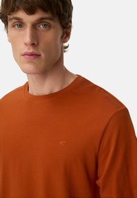 Ung mand med lys hud og kort krøllet brunt hår iført en simpel brændt orange T-shirt med rund hals mod en neutral baggrund.