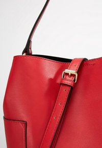 Červená kožená kabelka s hladkým povrchem, vybavená odnímatelným popruhem se zlatou sponou a černými dekoracemi uvnitř.
