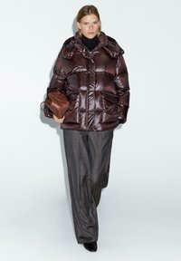 Donna che cammina indossando un piumino lucido di color maroon scuro, pantaloni grigi a gamba larga, scarpe nere e che tiene in mano una borsa marrone.