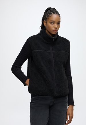 Vrouw met gevlochten haar, die een zwart fleecevest met rits draagt over een zwart shirt met lange mouwen en een donkere broek, staand met één hand in de zak.