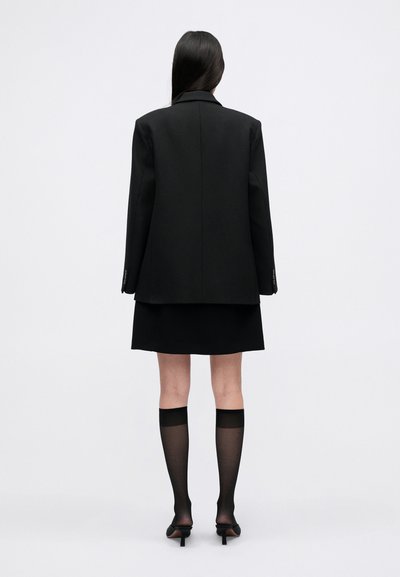 Blazer oversized preto com ombros estruturados, combinado com uma mini saia preta. Os acessórios incluem meias pretas transparentes e stiletto de bico fino.