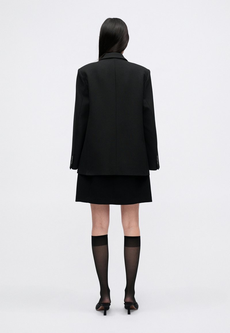 Black oversized blazer with structured shoulders, paired with a black mini skirt. Accessories include sheer black socks and pointed stilettos.
