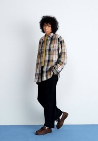 Camisa de cuadros en beige, verde y rojo con mangas largas, bolsillo en el pecho, combinada con una blusa verde y pantalones negros, y zapatos marrones.