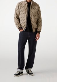 Uomo che indossa una giacca beige con motivo e zip sopra una maglietta nera, jeans scuri con risvolto e sneakers nere con lacci e suole bianche.