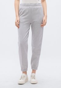 Pelēkas kokvilnas sporta bikses ar elastīgu vidukļa jostu, manžetēm pie potītēm un brīvu piegriezumu, kopā ar baltām sporta kurpēm.