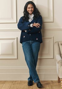 Námornícky pletený kardigán s kvetinovou výšivkou, v kombinácii s modrými džínsami a čiernymi topánkami. Stojí oproti textúrovanej stene na drevenej podlahe.