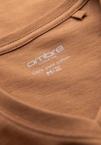 Collo di t-shirt in cotone marrone con una texture morbida. L'etichetta mostra il marchio "ombre", "100% cotone puro" e la taglia "M" (EUR 48).
