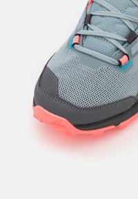 Zapatilla deportiva de malla gris con una superficie texturizada, detalles en rosa y suela de goma negra con trampas salientes. Cordones en azul y naranja.