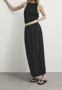 Haut cropped noir tricoté avec un design sans manches associé à une longue jupe noire plissée taille haute et des sandales plates noires.