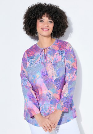 Kvinde med krøllet hår, der smiler, iført hængende øreringe og en langærmet bluse med flerfarvet mønster, stående foran en hvid baggrund.