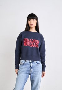 Sudadera navy con un gran texto gráfico rojo "NØRGAARD" exhibido de manera prominente. Presenta un corte relajado y puños acanalados. Combinada con jeans azul claro.