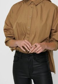 Brązowa bawełniana koszula zapinana na guziki z wywiniętym kołnierzykiem, zestawiona z czarnymi skórzanymi spodniami. Dłonie ozdobione wieloma cienkimi pierścieniami.