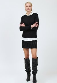 Pullover nero a maglia con orlo a coste, indossato sopra una camicia bianca con colletto e una gonna nera a righe verticali. Stivali neri in pelle al ginocchio.