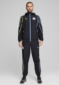 Tuta sportiva nera con giacca con cappuccio e cerniera e pantaloni abbinati. Accentuata da strisce blu chiaro e giallo neon. Logo e distintivo Puma visibili.