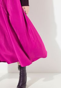 Pitkä, valuva mekko kirkkaan fuksia-kankaasta, joka on sileä tekstuuriltaan, yhdistettynä tiukkaan, tumman violetin nahkasaappaan.
