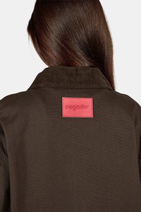 Pruunist korduroožakk, millel on silmapaistev krae ja roosa reljeefne silt seljal, esitleb tekstureeritud kangast ja puhtaid jooni.