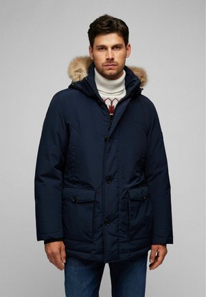 Navyblauwe gewatteerde parka met een met bont gevoerde capuchon, knopen aan de voorkant en twee buitenzakken. Draag het over een witte coltrui met een patroon.