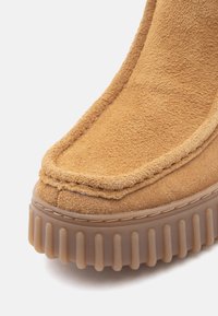 Tan mockasiner i mocka med rund tå, kontrasterande sömmar och en tjock genomskinlig gummisula med vertikala räfflor.