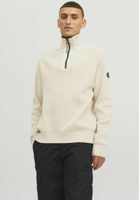 Beige gebreide pullover met een hoge kraag, rits tot de halve hoogte en geribbelde manchetten. Bevat een subtiele logopatch op de linkermouw.