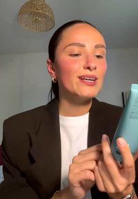 Frau mit dunklem Haar in einem braunen Blazer liest und zeigt auf eine blaue Tube, mit einer geflochtenen Lampe, die in einem schlichten Raum darüber hängt.