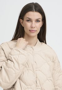 Jachetă bej matlasată cu decolteu rotund, având un model texturat, mâneci lungi și închidere frontală cu capse.