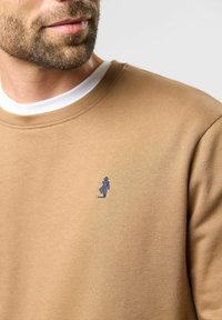 Ruskea collegepaita, jossa on pyöreä kaula-aukko ja vasemmassa rinnassa sininen brodé-merkki. Sileä rakenne, minimalistinen muotoilu, ilman kuvioita.
