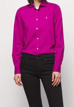 Persoon draagt een felroze blouse met lange mouwen, knoopsluiting en een klein geborduurd logo, gecombineerd met zwarte strakke broek.