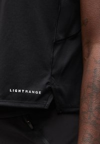 Κοντινό πλάνο μαύρου υφασμάτινου πουκαμίσου με υφή και το κείμενο "LIGHTRANGE" κοντά στο στρίφωμα, με ένα χέρι με τατουάζ δίπλα.