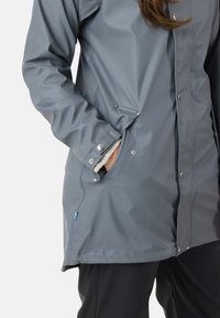 Personne portant une veste imperméable grise à boutons-pression, une main dans la poche latérale, et un pantalon noir visible sous la veste.