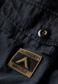 Camisa de botones color marino con un parche bordado del logo de Superdry en marrón. Presenta un cuello, un bolsillo lateral y una tela lisa y texturizada.