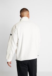 Mężczyzna z krótkimi włosami, ubrany w biały polar i czarne spodnie, stoi tyłem na jednolitym szarym tle.