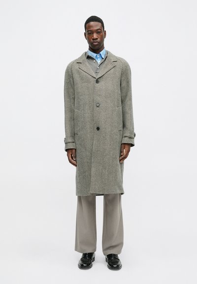 Sivi kaput od riblje kosti s kopčanjem na dugme, ravnim ovratnikom i dva bočna džepa, uparen s svijetlosivim hlačama i crnim cipelama.