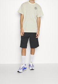 Randig ljusgul T-shirt med kort ärm, parat med svarta shorts, vita strumpor och blå och vita sneakers.