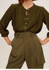 Blusa verde oliva con scollo rotondo, bottoni frontali e maniche a sbuffo abbinata a pantaloni verde oliva a vita alta con tasche piatte.