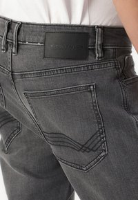 Jeans de mezclilla gris con una etiqueta de cuero negro en la cintura. Presentan costuras en contraste, bolsillos estándar y un acabado texturizado.