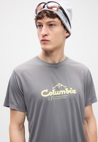 Γκρι αθλητική μπλούζα με κοντά μανίκια που διαθέτει κίτρινο λογότυπο "Columbia" με γραφικό βουνό, ομαλή υφή και επίπεδα ραφές.