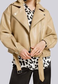 Giubbotto biker corto in ecopelle beige con chiusura a zip, colletto oversize e vita con cintura. Sotto c'è una blusa a fantasia bianco e nero.