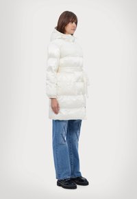Hvit pufferjakke med hette, knappelukking og belte i midjen. Kombinert med blå jeans med logomønster og svarte, tykke sko.