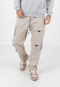 Beige Cargo-Hosen mit mehreren Seitentaschen, elastischem Bund und Reißverschlüssen. Kombiniert mit beigen und grauen Sneakers.