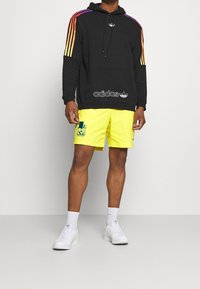 Black hoodie with multicolored stripes, front pocket, and logo. Yellow shorts with graphic detail. White shoes and socks worn.